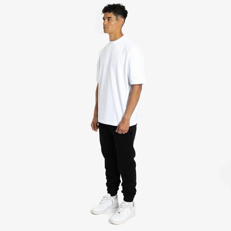 Puede incluir: Una persona que lleva una camiseta blanca de cuello redondo, pantalones de ch&aacute;ndal negros y zapatillas blancas. La camiseta tiene mangas cortas. Los pantalones de ch&aacute;ndal tienen cintura con cord&oacute;n y tobillos con pu&ntilde;os. Las zapatillas son bajas y con cordones.