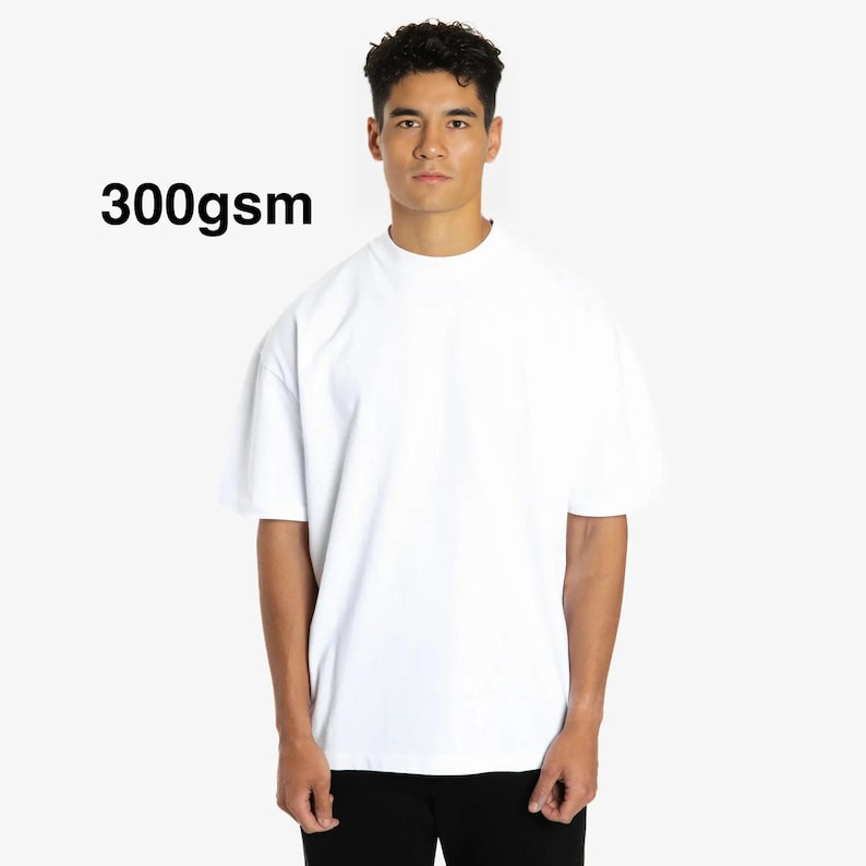 Puede incluir: Una camiseta blanca de gran tama&ntilde;o con cuello alto. El texto "300gsm" est&aacute; impreso en negro en la parte superior izquierda. La camiseta la lleva una persona con cabello oscuro y se combina con pantalones negros.