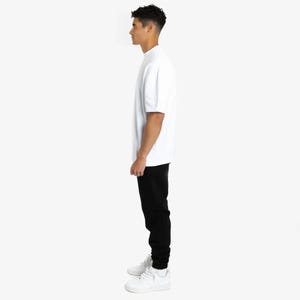 Puede incluir: Una persona vestida con una camiseta blanca de manga corta y pantalones negros. La persona tambi&eacute;n lleva zapatillas blancas. La camiseta tiene cuello redondo. Los pantalones son de color negro s&oacute;lido.