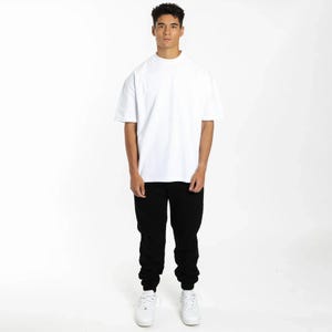 Puede incluir: Una persona que lleva una camiseta blanca de manga corta y un pantal&oacute;n de ch&aacute;ndal negro. La camiseta tiene cuello redondo. El pantal&oacute;n de ch&aacute;ndal tiene cintura con cord&oacute;n y tobillos con pu&ntilde;os. Zapatillas blancas completan el atuendo. El fondo es blanco liso.