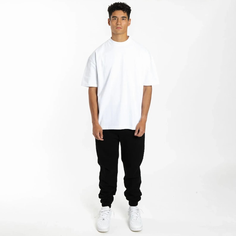 Puede incluir: Una persona que lleva una camiseta blanca de manga corta y un pantal&oacute;n de ch&aacute;ndal negro. La camiseta tiene cuello redondo. El pantal&oacute;n de ch&aacute;ndal tiene cintura con cord&oacute;n y tobillos con pu&ntilde;os. Zapatillas blancas completan el atuendo. El fondo es blanco liso.