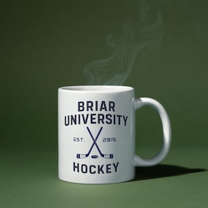 Op de afbeelding: Een witte keramische mok met de tekst "BRIAR UNIVERSITY HOCKEY" in marineblauw. De mok heeft gekruiste hockeysticks en het oprichtingsjaar "2015". Stoom stijgt op uit de mok tegen een groene achtergrond.