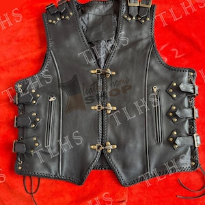 Peut inclure: Gilet en cuir noir avec un design tressé, de multiples boucles et des clous dorés. Le gilet a deux poches zippées et un dos à lacets.
