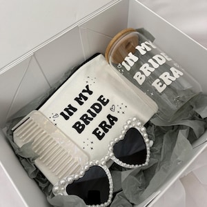 Könnte beinhalten: Eine weiße Geschenkbox enthält eine Kosmetiktasche, eine Sonnenbrille, einen Kamm und einen Glasbecher. Die Kosmetiktasche und der Becher tragen den Text "IN MY BRIDE ERA". Die Sonnenbrille hat einen herzförmigen Rahmen mit Perlenakzenten.
