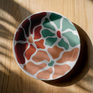 Puede incluir: Un plato redondo de cer&aacute;mica blanca con un colorido dise&ntilde;o floral abstracto. El dise&ntilde;o presenta formas org&aacute;nicas en tonos de rojo, naranja y verde. El plato est&aacute; sobre una superficie de madera.