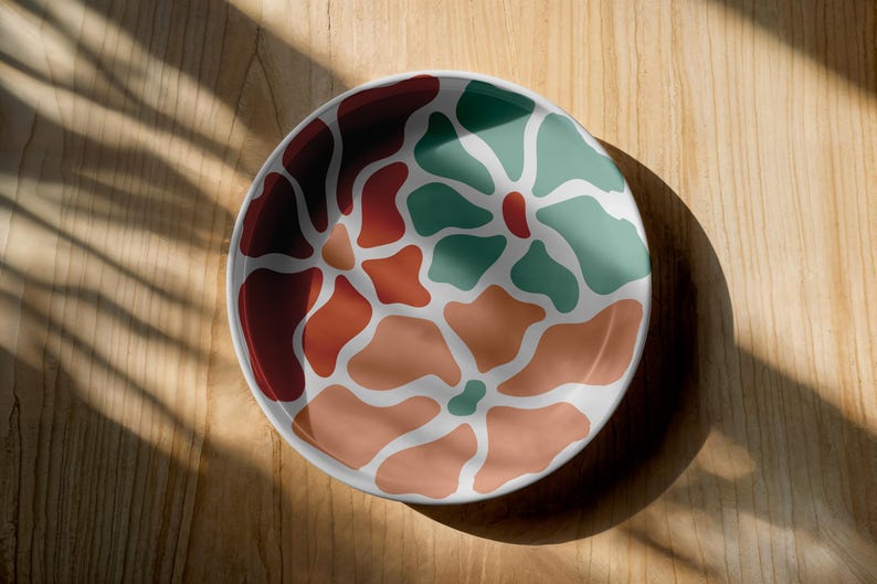 Puede incluir: Un plato redondo de cer&aacute;mica blanca con un colorido dise&ntilde;o floral abstracto. El dise&ntilde;o presenta formas org&aacute;nicas en tonos de rojo, naranja y verde. El plato est&aacute; sobre una superficie de madera.
