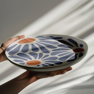 May include: A white ceramic serving platter with a blue and orange floral design. The platter is being held by a hand.