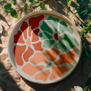 Puede incluir: Una bandeja decorativa redonda de color crema con un dise&ntilde;o floral colorido. El dise&ntilde;o presenta formas abstractas en tonos de rojo, naranja y verde. La bandeja est&aacute; rodeada de hojas y vides verdes.