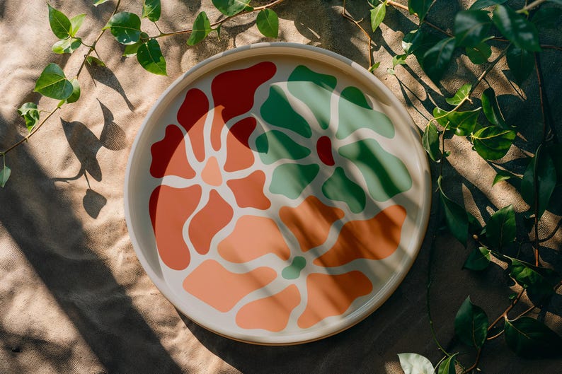 Puede incluir: Una bandeja decorativa redonda de color crema con un dise&ntilde;o floral colorido. El dise&ntilde;o presenta formas abstractas en tonos de rojo, naranja y verde. La bandeja est&aacute; rodeada de hojas y vides verdes.