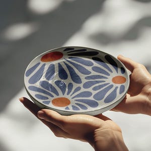 May include: A white ceramic bowl with a blue and orange floral design. The bowl is being held by a person's hands.