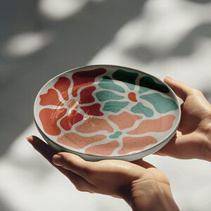 Puede incluir: Un plato de cer&aacute;mica con un dise&ntilde;o abstracto colorido. El plato presenta un borde blanco y un patr&oacute;n de formas rojas, coral y turquesa. El plato est&aacute; sostenido en dos manos.