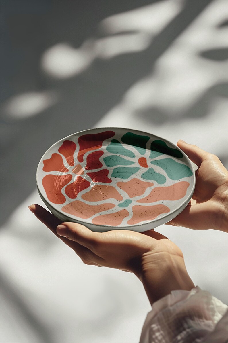 Puede incluir: Un plato de cer&aacute;mica con un dise&ntilde;o abstracto colorido. El plato presenta un borde blanco y un patr&oacute;n de formas rojas, coral y turquesa. El plato est&aacute; sostenido en dos manos.