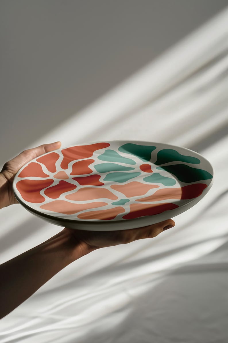 Puede incluir: Plato decorativo de cer&aacute;mica con dise&ntilde;o abstracto colorido. El plato presenta una base blanca con un patr&oacute;n de formas rojas, coral y turquesas. Se muestra sostenido por dos manos, resaltando su dise&ntilde;o.