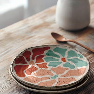 Puede incluir: Dos platos de cer&aacute;mica apilados sobre una mesa de madera. Los platos tienen una base blanca con un dise&ntilde;o abstracto colorido en tonos naranja, turquesa y melocot&oacute;n. Un jarr&oacute;n peque&ntilde;o con flores blancas y una cuchara de madera est&aacute;n en el fondo.