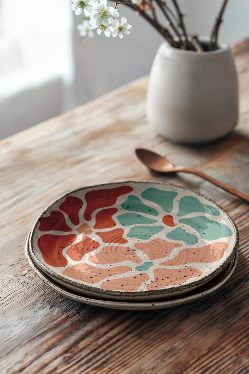 Puede incluir: Dos platos de cer&aacute;mica apilados sobre una mesa de madera. Los platos tienen una base blanca con un dise&ntilde;o abstracto colorido en tonos naranja, turquesa y melocot&oacute;n. Un jarr&oacute;n peque&ntilde;o con flores blancas y una cuchara de madera est&aacute;n en el fondo.