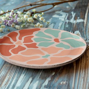 Puede incluir: Un plato decorativo de cer&aacute;mica con un dise&ntilde;o floral en tonos coral y turquesa. El plato se encuentra sobre una superficie de madera envejecida, te&ntilde;ida de azul, con una ramita de flores en el fondo.