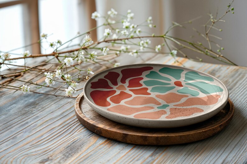 Puede incluir: Un plato de cer&aacute;mica con un dise&ntilde;o floral en tonos rojos, coral y verde azulado. El plato est&aacute; sobre una bandeja redonda de madera, con una ramita de flores blancas en el fondo. El plato tiene aproximadamente 30 cm de di&aacute;metro.