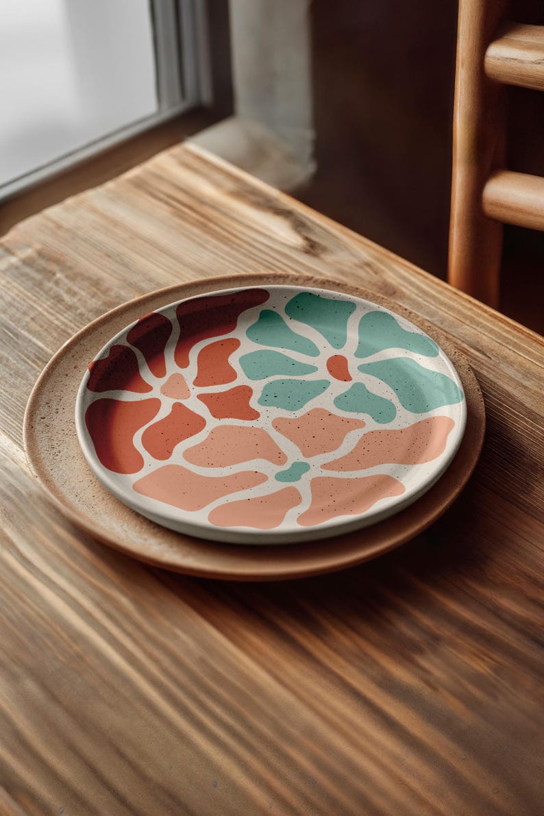 Puede incluir: Plato de cer&aacute;mica decorativo con un dise&ntilde;o floral en tonos coral, melocot&oacute;n y turquesa. El plato est&aacute; colocado sobre otro plato m&aacute;s grande, de tono neutro, creando un efecto de capas. El dise&ntilde;o presenta formas org&aacute;nicas y una textura moteada.