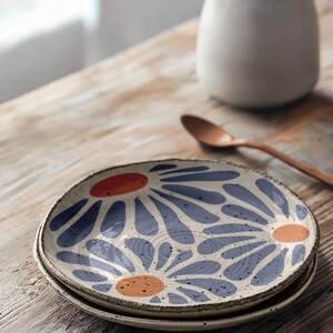 May include: A set of two ceramic plates with a white background and a blue and orange floral pattern. The plates are stacked on top of each other on a wooden table.