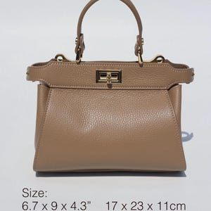 K&ouml;nnte beinhalten: Eine hellbraune Ledertasche mit goldfarbenem Verschluss und einem oberen Henkel. Die Tasche misst 17 x 23 x 11 cm.