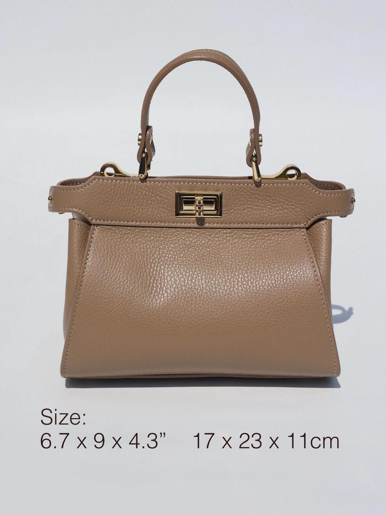 K&ouml;nnte beinhalten: Eine hellbraune Ledertasche mit goldfarbenem Verschluss und einem oberen Henkel. Die Tasche misst 17 x 23 x 11 cm.