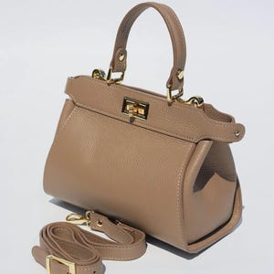 K&ouml;nnte beinhalten: Eine hellbraune Ledertasche mit einem Henkel oben und einem abnehmbaren Schultergurt. Die Tasche hat ein goldfarbenes Schloss und Beschl&auml;ge.