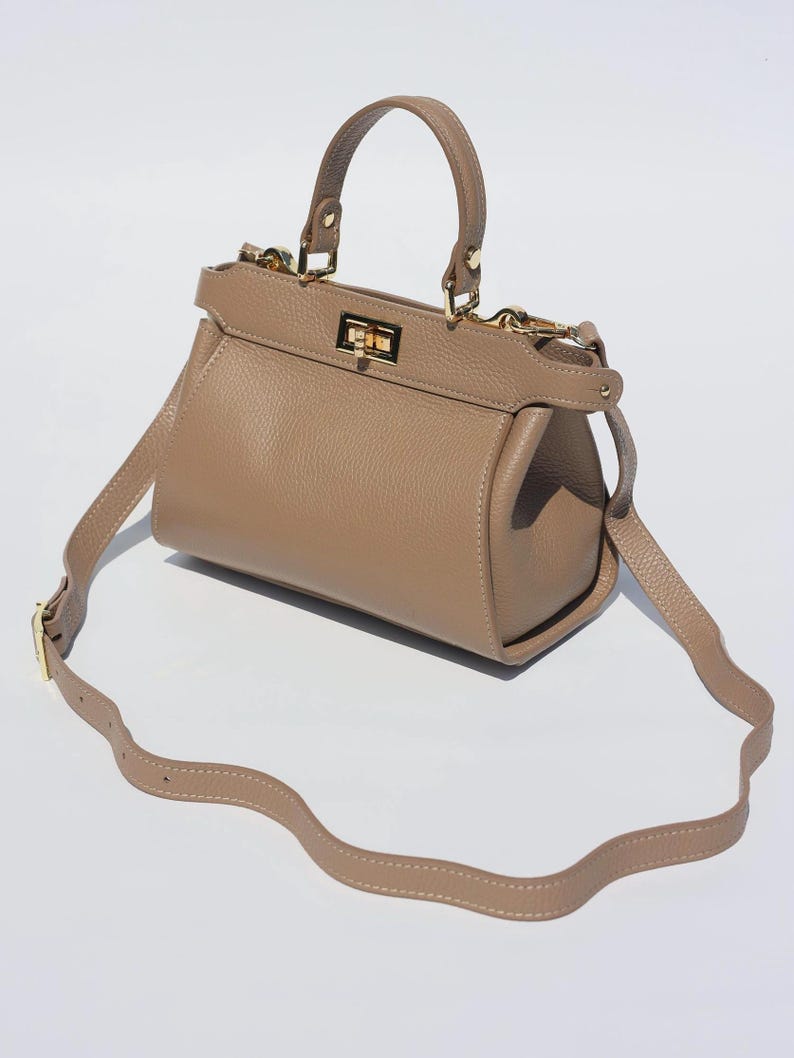 K&ouml;nnte beinhalten: Eine hellbraune Ledertasche mit einem Henkel oben und einem langen, verstellbaren Schultergurt. Die Tasche hat einen goldfarbenen Verschluss und Beschl&auml;ge.