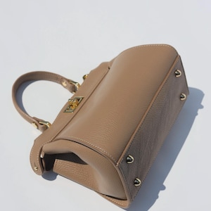 K&ouml;nnte beinhalten: Eine hellbraune Ledertasche mit goldenem Verschluss und goldenen Nieten. Die Tasche hat einen oberen Henkel und einen Klappenverschluss.