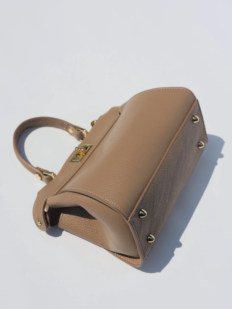 K&ouml;nnte beinhalten: Eine hellbraune Ledertasche mit goldenem Verschluss und goldenen Nieten. Die Tasche hat einen oberen Henkel und einen Klappenverschluss.