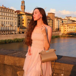 K&ouml;nnte beinhalten: Eine Frau in einem rosa T&uuml;llkleid mit einer braunen Ledertasche steht auf einer Steinbr&uuml;cke und blickt auf einen Fluss. Die Stadt Florenz in Italien ist im Hintergrund zu sehen.