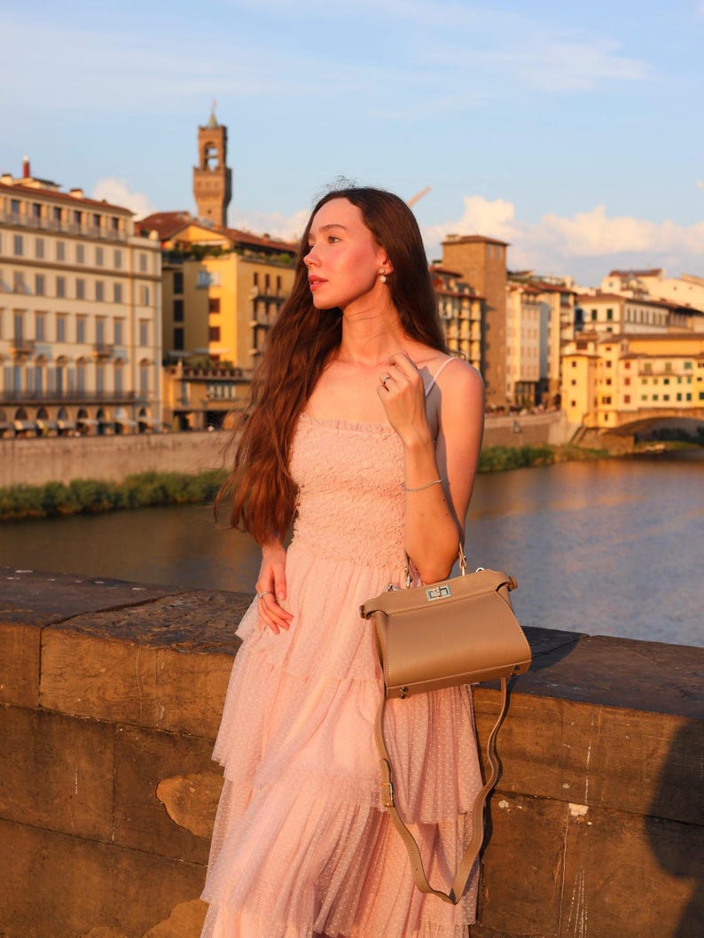 K&ouml;nnte beinhalten: Eine Frau in einem rosa T&uuml;llkleid mit einer braunen Ledertasche steht auf einer Steinbr&uuml;cke und blickt auf einen Fluss. Die Stadt Florenz in Italien ist im Hintergrund zu sehen.
