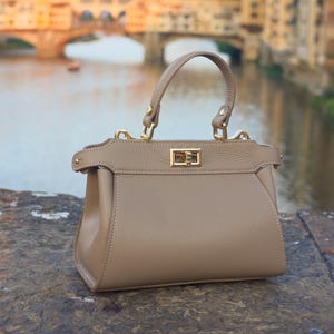 K&ouml;nnte beinhalten: Eine hellbraune Ledertasche mit einem Henkel oben und einem goldenen Verschluss. Die Tasche liegt auf einer Steinoberfl&auml;che mit einem verschwommenen Hintergrund einer Br&uuml;cke.