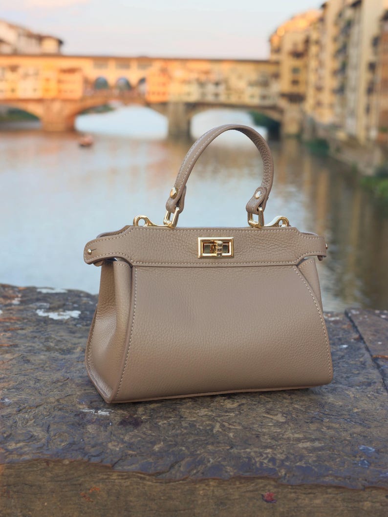 K&ouml;nnte beinhalten: Eine hellbraune Ledertasche mit einem Henkel oben und einem goldenen Verschluss. Die Tasche liegt auf einer Steinoberfl&auml;che mit einem verschwommenen Hintergrund einer Br&uuml;cke.