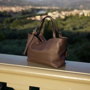 May include: A brown leather tote bag with two handles and a shoulder strap. The bag has a textured surface and is resting on a white railing. The background shows a blurred view of a city and trees.