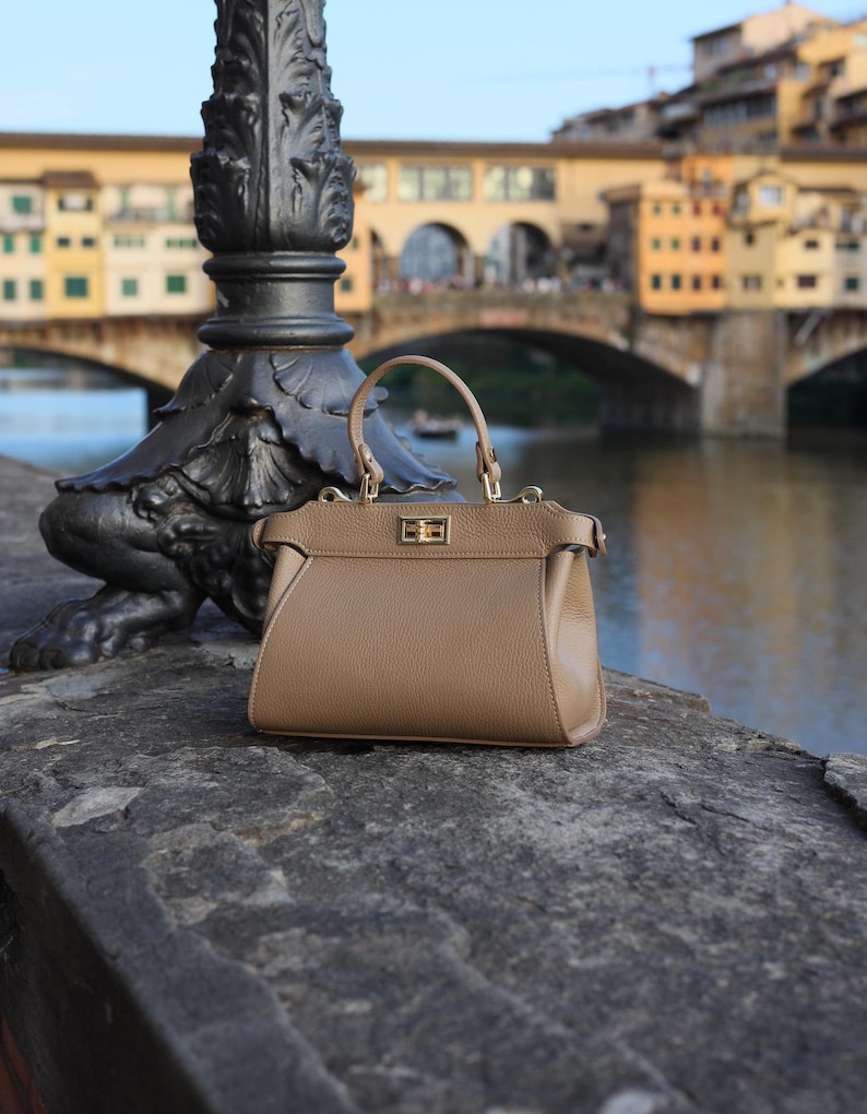 K&ouml;nnte beinhalten: Eine braune Ledertasche mit goldenem Verschluss und einem oberen Henkel. Die Tasche liegt auf einer Steinoberfl&auml;che vor einer Br&uuml;cke.