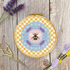 Può includere: Un telaio da ricamo a punto croce con un motivo a forma di ape e dettagli di lavanda. Il disegno è ambientato su uno sfondo azzurro con un bordo a scacchi giallo e bianco. Sono presenti anche forbici dorate e filo da ricamo.
