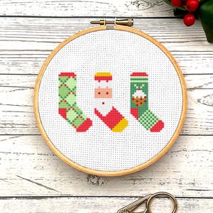 Peut inclure: Un gros plan d'un cerceau de broderie en point de croix avec trois chaussettes de Noël. Les chaussettes sont vertes et rouges avec un fond blanc. Une chaussette a un motif de Père Noël, une autre a un motif de pudding de Noël et la troisième a un motif à carreaux.
