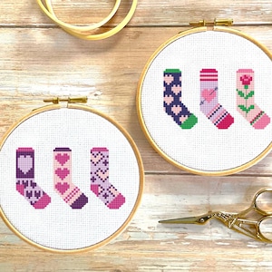 Peut inclure: Deux cerceaux à broder avec un tissu blanc et des motifs de point de croix colorés de chaussettes avec des cœurs, des flèches et des fleurs. Les cerceaux sont sur une surface en bois avec une paire de ciseaux dorés.