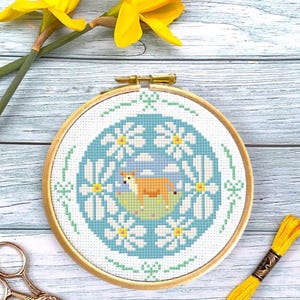 May include: A cross-stitch embroidery hoop with a blue background and a white daisy wreath. A brown cow is in the center of the wreath, standing on a green field.