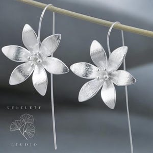 Op de afbeelding: Een paar zilveren bloemvormige oorhangers met een textuur. De oorhangers hangen aan een dunne zilveren draad.