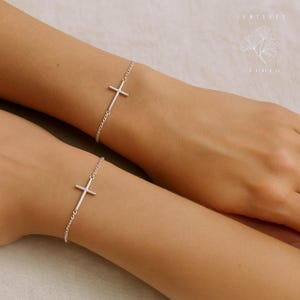 May include: Two silver-colored cross bracelets are displayed on wrists. The bracelets feature a simple cross design and are connected by a delicate chain. The background is a soft, neutral color, highlighting the jewelry.