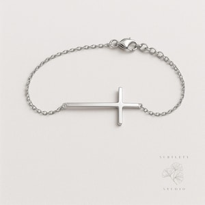 Könnte beinhalten: Ein silbernes Armband mit einem seitlichen Kreuzanhänger. Das Kreuz ist mit einer feinen Kette mit einem Karabinerverschluss verbunden. Dieses elegante Armband ist ein schlichtes Schmuckstück.