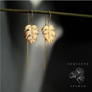 Pode incluir: Brincos de folha de ouro com um delicado design de folha de monstera texturizada. Os brincos são longos e pendentes, com um fio fino de ouro que conecta a folha ao gancho.