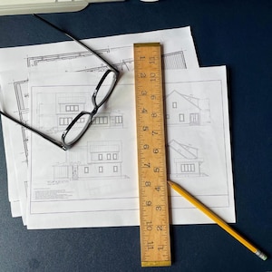 May include: Architectural blueprints with house plans, a wooden ruler measuring 11 inches, a yellow pencil, and black-framed glasses. The blueprints show detailed drawings of a house, including elevations and floor plans. The ruler is marked with inch measurements.