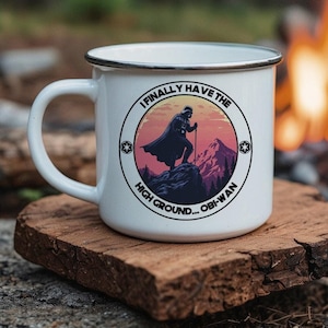 May include: White enamel camping mug with a black rim and handle. The mug features a graphic of a character standing on a mountain with the text "I FINALLY HAVE THE HIGH GROUND... OBI-WAN". The mug is sitting on a wooden surface near a campfire.