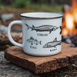 May include: White enamel camping mug featuring black illustrations of fish, including sturgeon, perch, and barbel. The mug is resting on a piece of wood near a campfire.