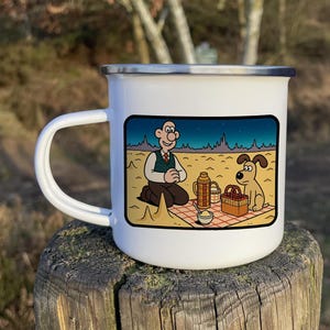 May include: White enamel camping mug featuring a cartoon illustration of a man and a dog enjoying a picnic in a desert setting. The mug has a black handle.