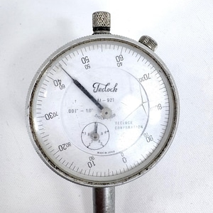 May include: A close-up of a Teclock dial indicator, model AI-921, with a white face and black markings. The dial measures from .001" to 1.0". The indicator has a silver metal casing and a stem.