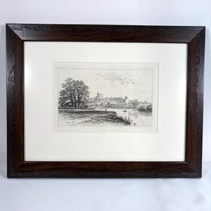 May include: A framed black and white landscape print featuring a detailed depiction of a town or city along a river. The print is set within a white mat and a dark brown wooden frame, creating a classic aesthetic. The artwork showcases trees, buildings, and a body of water.
