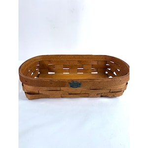 May include: A rectangular woven wooden basket with rounded corners and a flat base. The basket features a medium brown finish and a small, dark oval metal tag centered on the front rim with text that reads Longaberger 1997 Dresden Ohio.